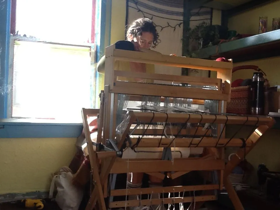 A woman sits at a loom weaving in the corner of a room next to a bright window.