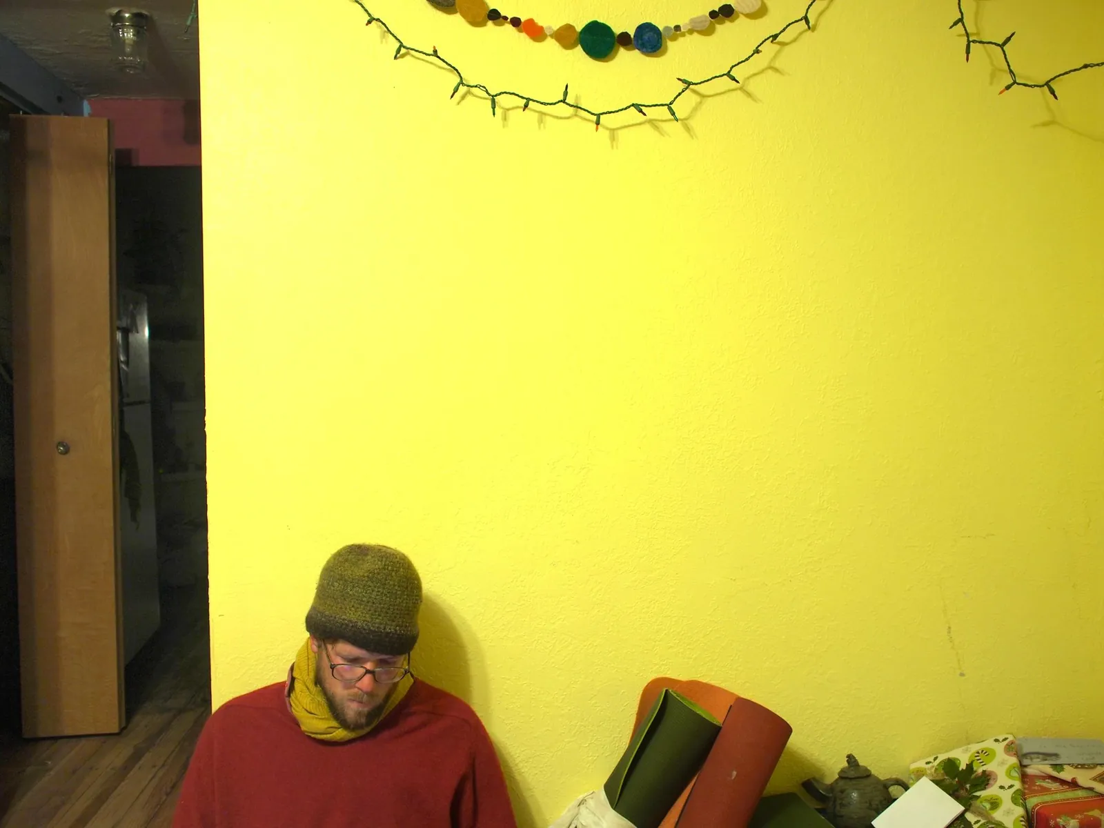 A yellow wall fills most of the picture while a man sitting on the floor looks down at his hands.