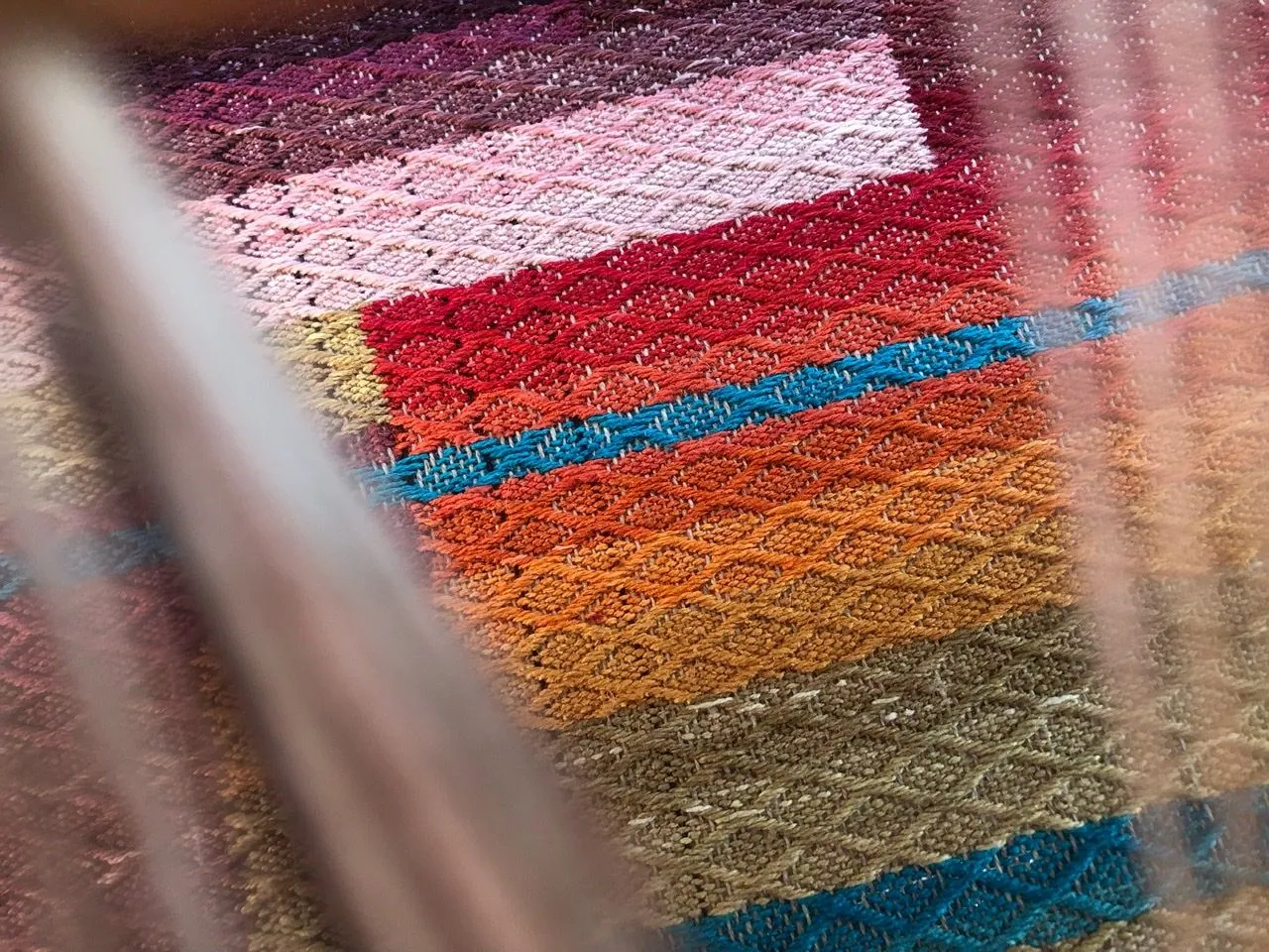 An up close image looking through the threads of a loom at a blocky, rainbow colored diamond pattern weaving