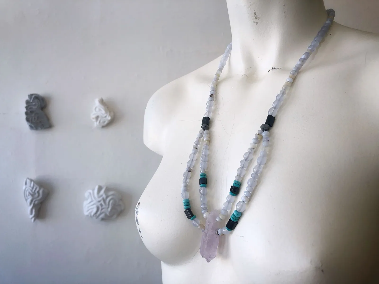 A white mannequin wears a necklace of pale blue, purple, turquoise and black semi precious stones in a gallery with marble sculptures on the wall