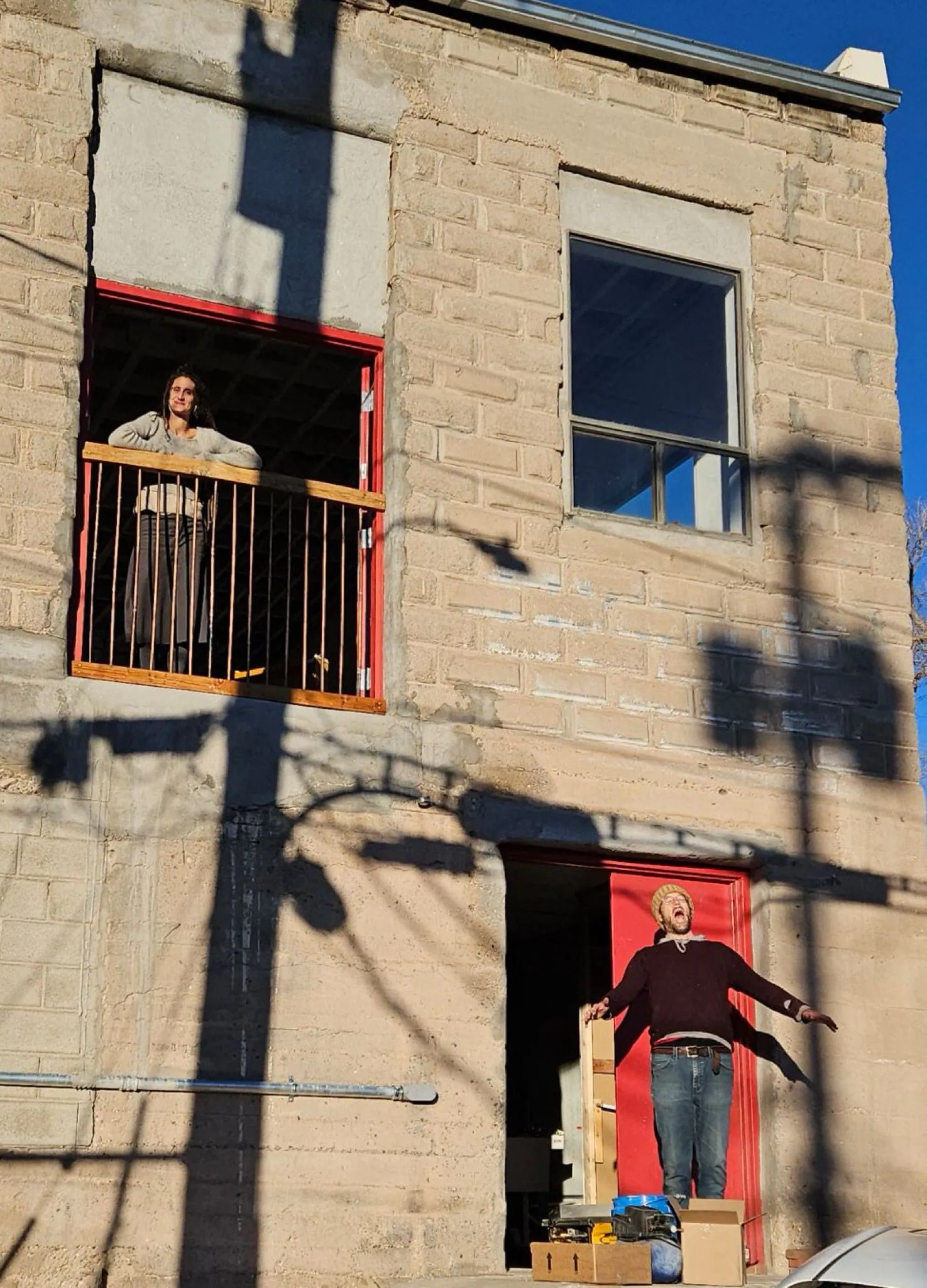 The large concrete exterior facade of a building with two red doors, one on ground level and one on a second story, a women is in the upper doorway, a man below.