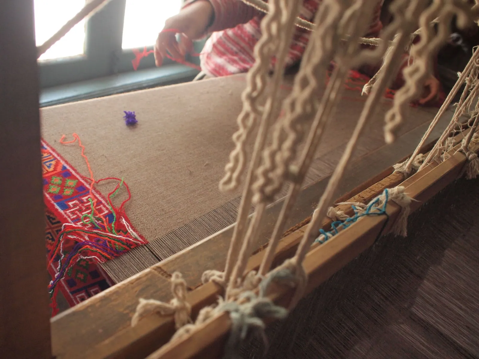 A persons hands working at loom, weaving a brown-tan blanket with a brightly colored red orange, blue, green and white geometric border.
