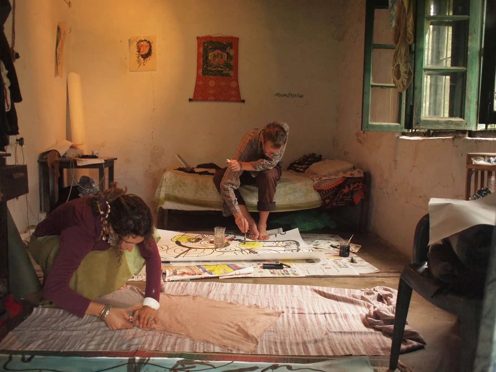 A woman wearing a raspberry shirt and green skirt squats on the floor, cutting out fabric for a dress. In the background a man sits on a small bed painting. There is a green trimmed window open to the right and a red bordered thangka on the wall behind.