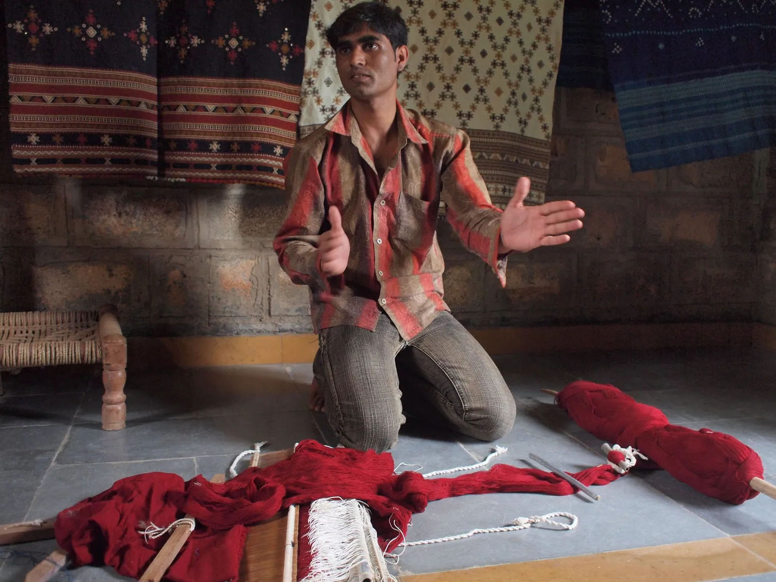 A man in red and brown shirt kneels on a grey stone floor, in front of him are pieces of a loom and a spool of bright red threads.