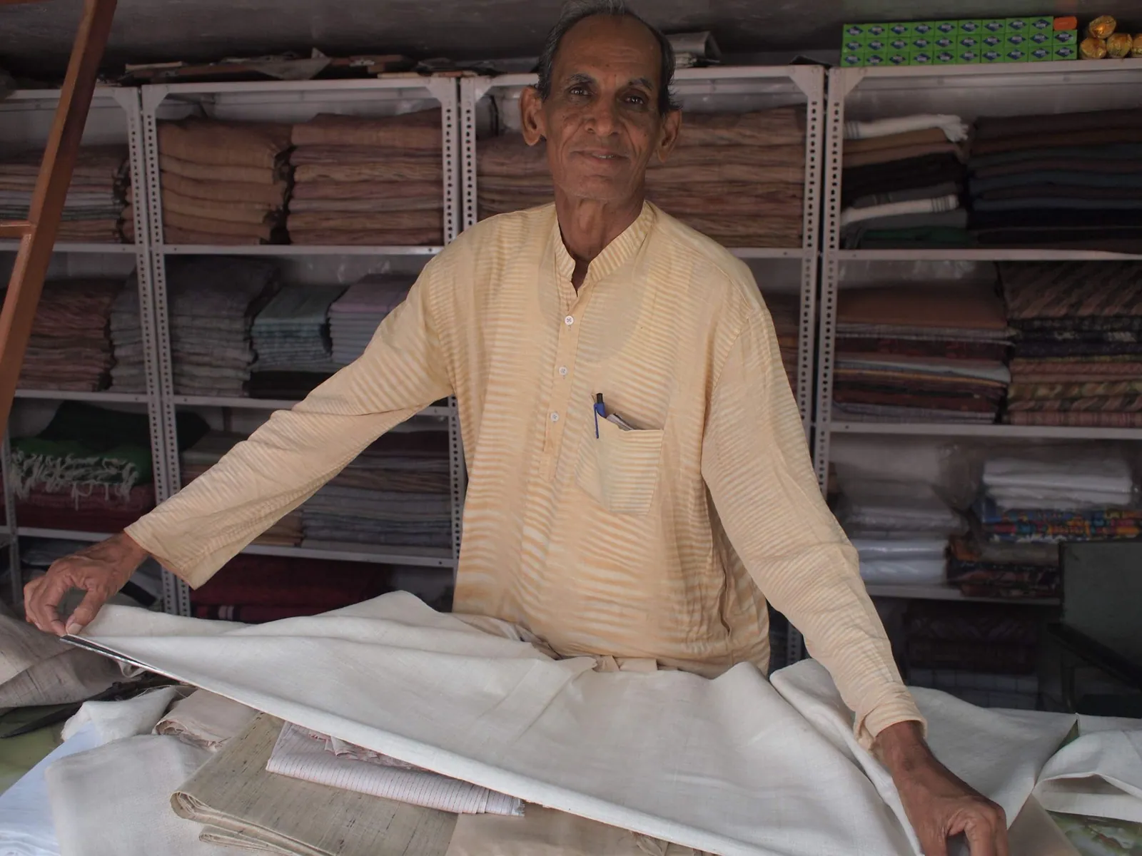a man in a pale yellow and white shirt measures out white fabric with a wall full of stacked fabric behind him.
