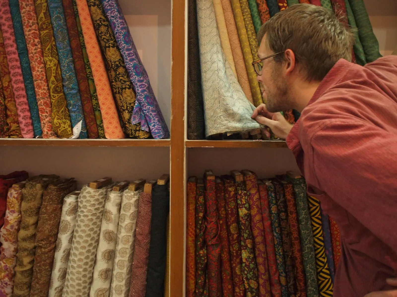 A man with glasses looks closely at white and blue fabric that is stacked on shelves of bolts of fabric of every color.