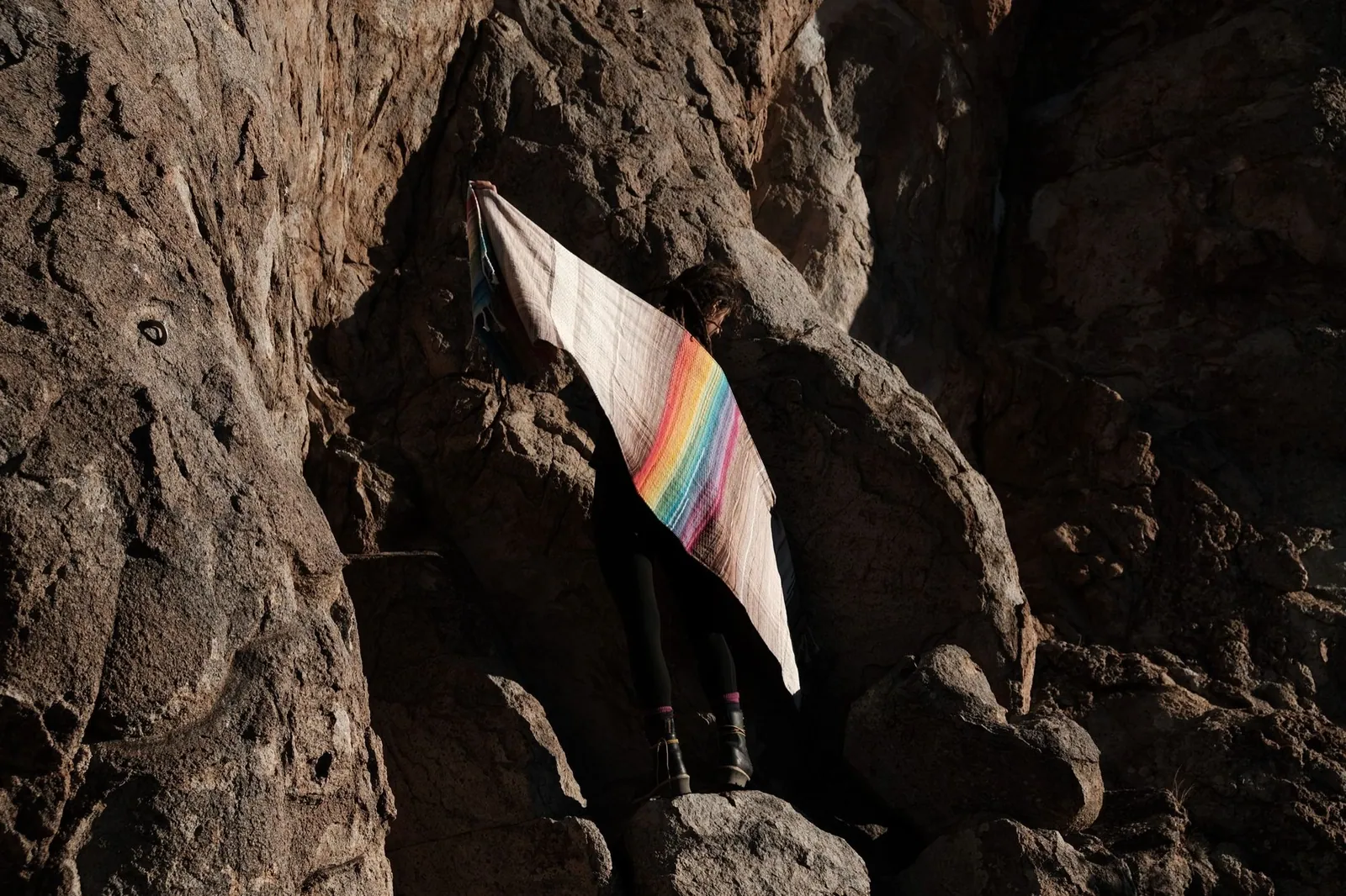 A women stands in the shadow of a granite cliff silhouetted by evening light while holding a rainbow and grey handwoven shawl.