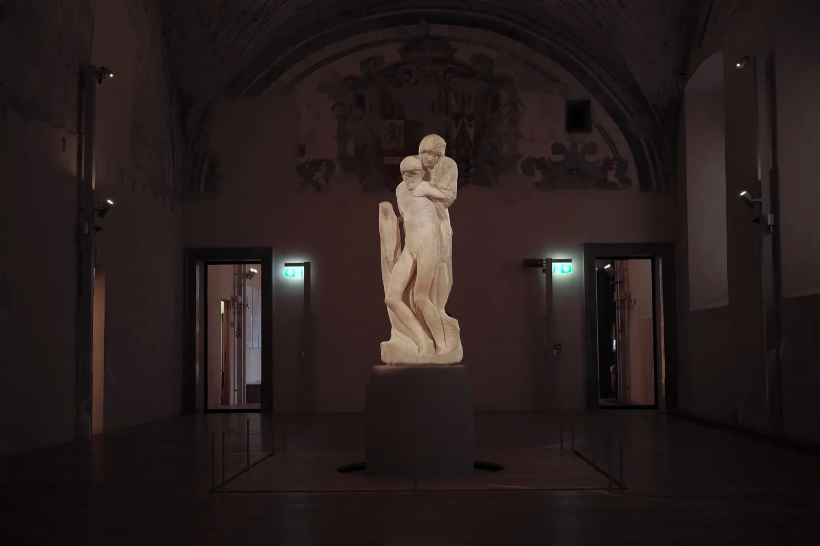 A sculpture of two figures, one holding the limp body of another, stands illuminated in center of a darkened room.