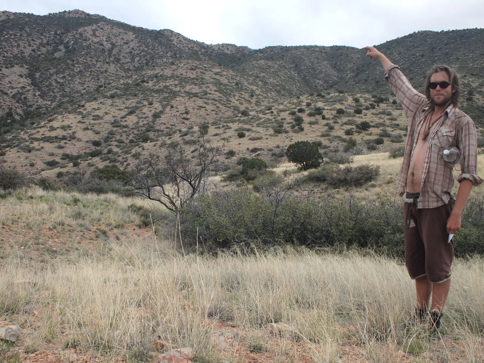 A man in plaid shirt and brown shorts stands in a desert landscape and points to the top of a mountain.