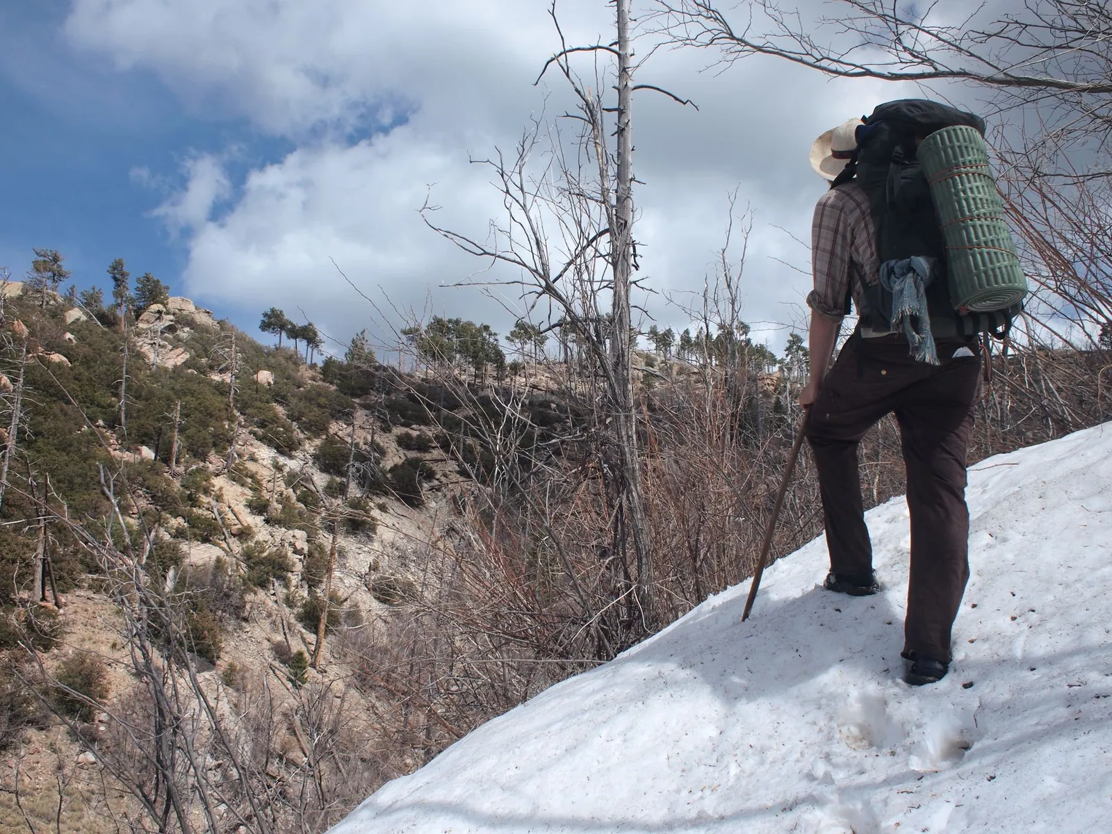 Man in plaid shirt and brown pants wearing large backpack, hiking through the snow in the mountains