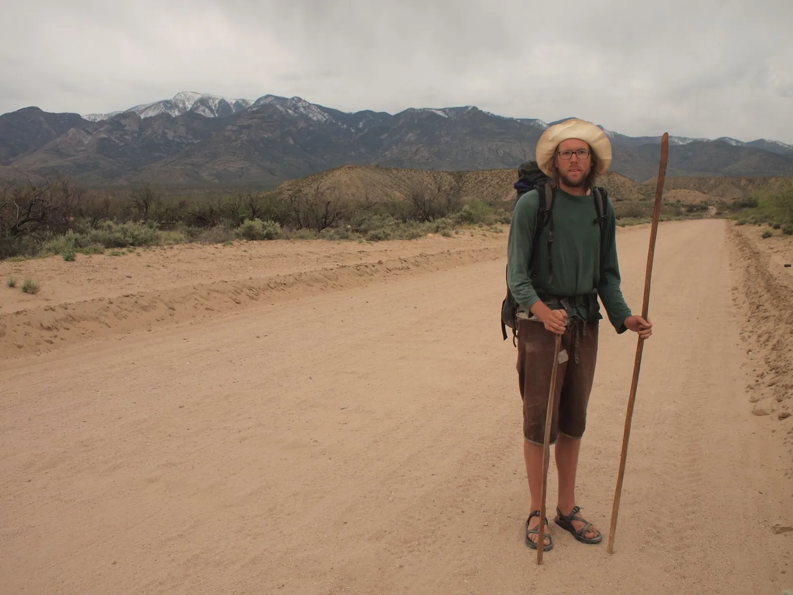 A man stands in the middle of a dirt road in the desert with snowy mountains behind.